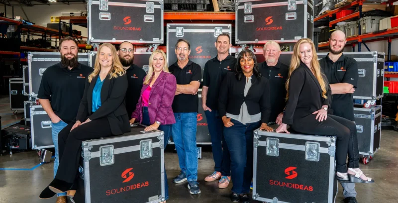 Corporate leadership team posing in warehouse with branded equipment cases, representing teamwork, trust, and organizational alignment