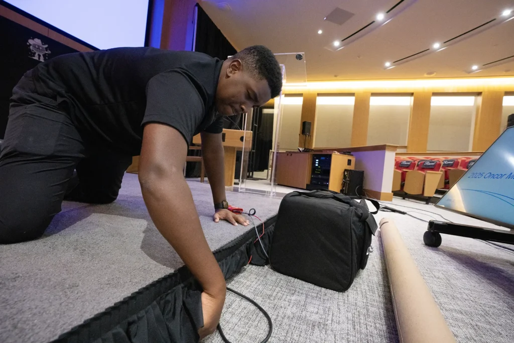 An AV technician routes cables beneath a conference stage beside presentation screens.