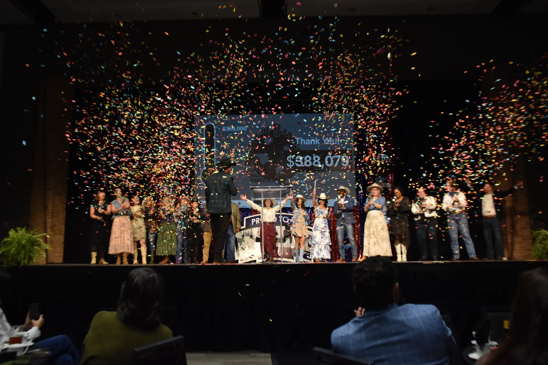 People on stage celebrate under colorful confetti with a fundraising total displayed behind them.