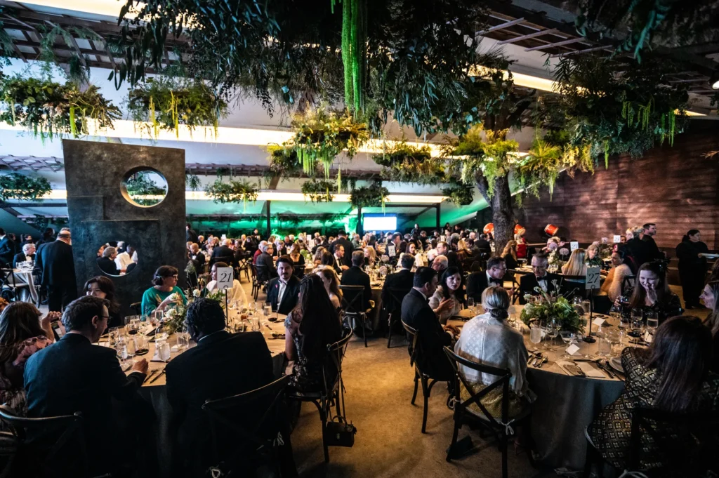 Guests in formal attire sit at decorated round tables under hanging greenery at a fundraising gala.