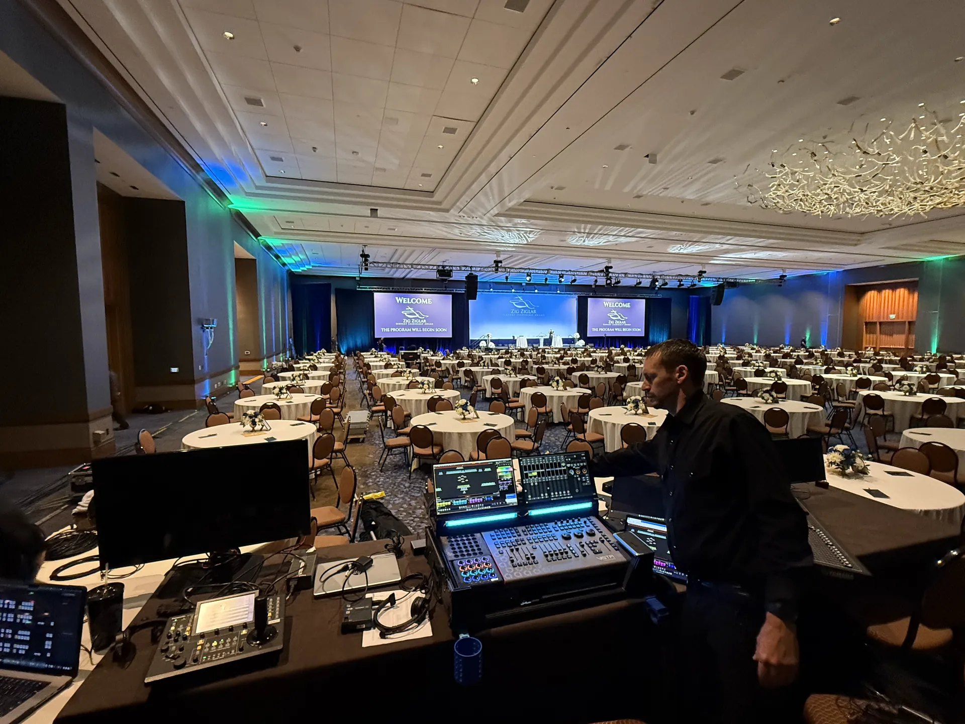 A man operates sound and lighting equipment at the back of a large empty gala event room.